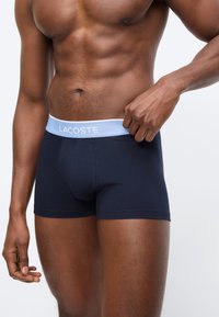 Μυώδες σώμα άνδρα που φοράει μπλε σκούρα εσώρουχα Lacoste με ανοιχτό μπλε ζώνη στη μέση, ρυθμίζοντας τη ζώνη με το δεξί χέρι.