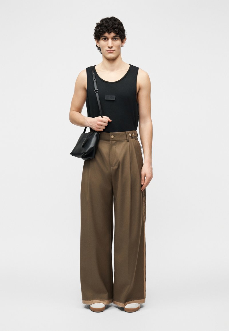 Young person wearing a black sleeveless top, wide brown pants, white shoes, and carrying a small black crossbody bag on a plain background.