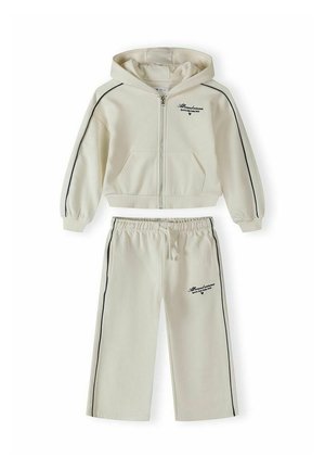 Dwuczęściowy kremowy dres z kapturem, rozpinaną na zamek bluzą oraz spodniami z elastyczną talią, obie części ozdobione czarnymi paskami po bokach i małymi logo z tekstem.