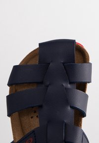 Tummansininen nahkasandaali, jossa on kolme vaakasuoraa hihnaa ruskealla mokkanahkapedillä ja punainen yksityiskohta kantapään lähellä.