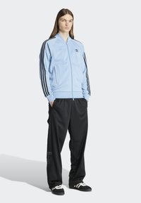 Světle modrá sportovní bunda na zip s černými pruhy, v kombinaci s černými kalhotami a černými botami s bílými akcenty. Materiál vypadá hladce.