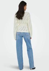 Pull crème en tricot avec un dos en V, un motif ajouré et un détail noué à la taille ; associé à un jean bleu clair taille haute.