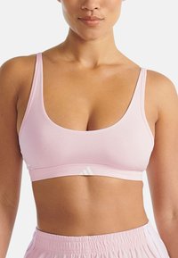 Jasnoróżowy stanik sportowy wykonany z gładkiego materiału, z okrągłym dekoltem i cienkimi ramiączkami. Zawiera biały akcent z logo na pasie.