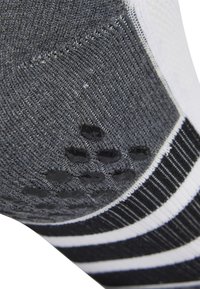 Calcetín deportivo gris con tela de punto texturizada, puntos de agarre de goma en la suela y rayas en blanco y negro en la parte superior.