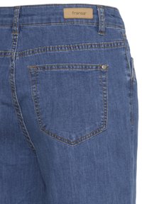 Gros plan sur l'arrière d'un jean en denim bleu avec une seule poche arrière et une étiquette beige portant l'inscription "fransa" à la ceinture.
