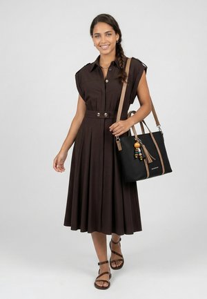 Frau mit geflochtenem Haar, die ein braunes ärmelloses knöpfbares Midikleid und Sandalen trägt, hält eine schwarze Handtasche mit braunen Trägern und dekorativen Perlen.