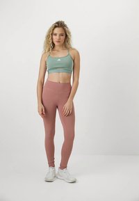 Femme debout en tenue de sport, portant un soutien-gorge de sport vert, des leggings roses et des baskets blanches sur un fond uni.