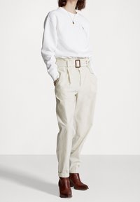 Kvinna i en vit långärmad skjorta, beige, högmidjade byxor med bälte, bruna ankelboots och ett runt hängsmycke.