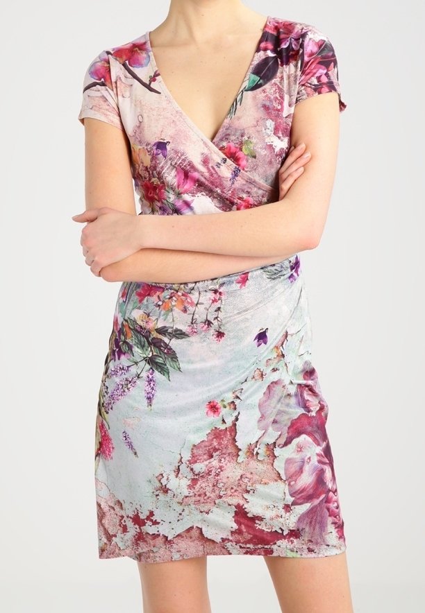 Femme portant une robe à manches courtes et col en V avec des motifs floraux roses, violets et verts sur un fond clair, les bras croisés.