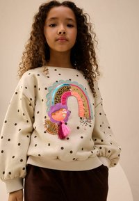 Cremefarvet sweatshirt med sorte prikker, prydet med et farverigt regnbue-design med dekorationer, lagdelte teksturer og en lyserød frynse.