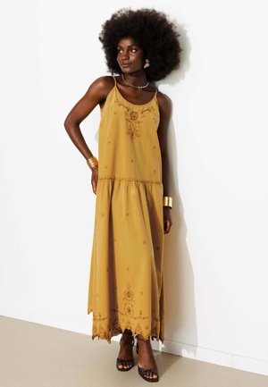 Femme avec coiffure afro porte une robe longue jaune moutarde à fines bretelles et des talons marron, accessoirisée de bijoux en or, debout contre un mur blanc.