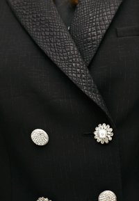 Czarna marynarka z fakturowanym czarnym satynowym kołnierzem, ozdobiona trzema dekoracyjnymi guzikami: perłą, kwiatowym wzorem oraz fakturowanym okrągłym kształtem.