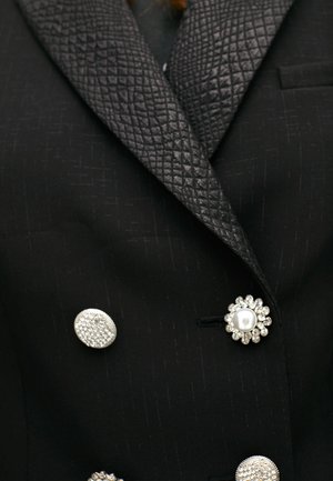Blazer nero con revers in satin nero testurizzato, decorato con tre bottoni ornamentali: una perla, un design floreale e una forma circolare testurizzata.