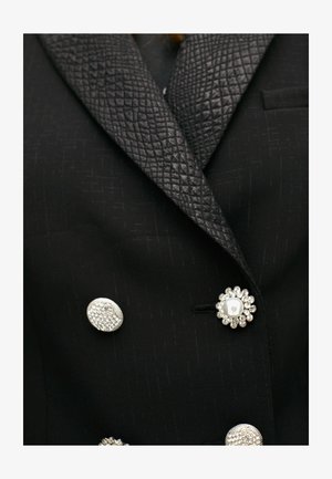 Schwarzer Blazer mit einem strukturierten schwarzen Satin-Revers, verziert mit drei dekorativen Knöpfen: einem Perlenknopf, einem Blumenmuster und einer strukturierten runden Form.