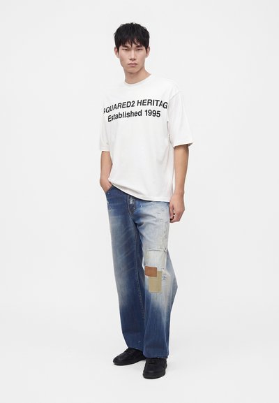 Νέος άνδρας φοράει λευκό φαρδύ T-shirt με μαύρο κείμενο, φαρδιά μπλε τζιν με patches και μαύρα παπούτσια, στέκεται σε λευκό φόντο.