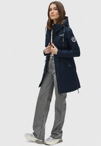 Navy waterproof jacket with adjustable hood and zippered pockets, paired with gray straight-leg jeans and light gray sneakers.