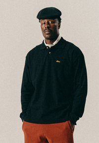 Man in een zwarte platte pet, zwarte longsleeve polo met gouden logo, een gestreepte nek sjaal en roestkleurige broek met handen in de zakken.