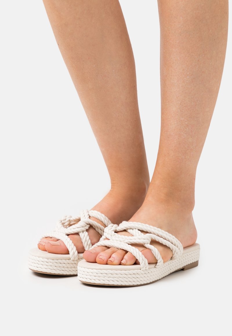 Pieds portant des sandales en corde blanches avec un motif de nœud croisé et des semelles plates tissées sur un fond uni.