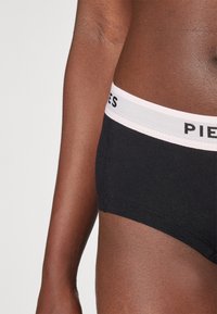 Crne pamučne gaćice s mekim elastičnim pojasom na kojem je u crnom slovima ispisano "PIECES" na svijetlo ružičastoj pozadini. Glatka tekstura.