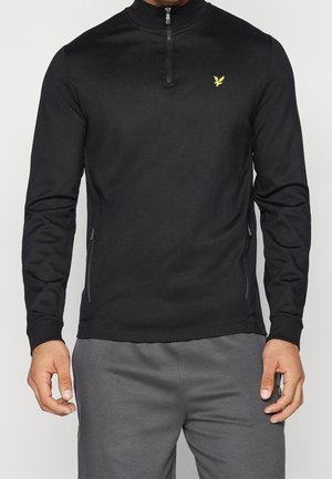 Mężczyzna ubrany w czarną bluzę z długim rękawem, zapinaną na zamek do połowy, z żółtym logo ptaka, w połączeniu z szarymi spodniami.