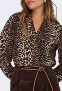 Femme portant un chemisier imprimé léopard avec un profond décolleté en V, rentré dans un pantalon en velours côtelé marron taille haute, maintenu par une fine ceinture marron.
