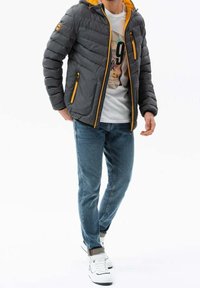 Chaqueta acolchada gris con capucha amarilla, dos bolsillos con cremallera y un diseño acolchado. Lleva puesta una camiseta gráfica blanca y jeans azules.