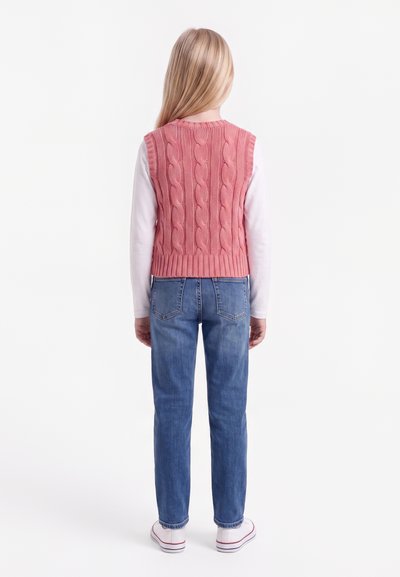 Enfant aux longs cheveux blonds portant un gilet en maille rose sur une chemise blanche à manches longues et un jean bleu, debout de dos.