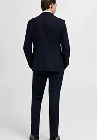Marineblå jakkesæt med en skræddersyet blazer og bukser, der har en glat tekstur og en enkelt slids bagpå, parret med sorte sko.