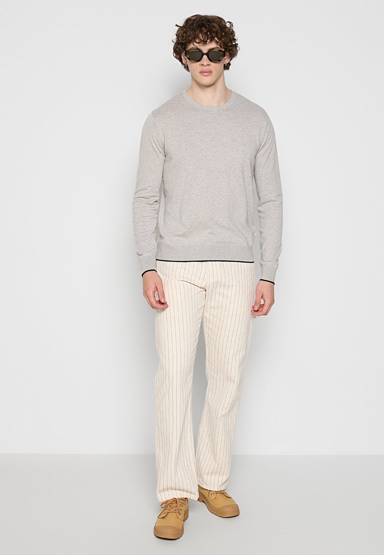 Grauer langärmeliger Pullover, cremefarbene gestreifte Hose und beigefarbene Schuhe. Der Pullover hat einen runden Ausschnitt; die Hose hat vertikale Streifen.