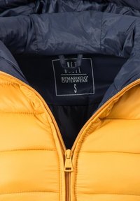 Gul pufferjakke med en marineblå indvendig side, med quiltet tekstur og lynlås foran. Mærket læser "BYMARIKOO OUTERWEAR," størrelse S.