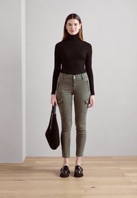 Črn pulover z visoko ovratnikom iz pletenine, olivno zelene hlače s predali in zadrgami, črna torba in črne platformne loafers.