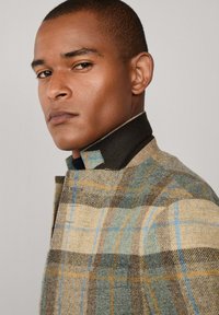 Chaqueta de lana a cuadros en tonos terrosos de beige, marrón, verde y azul, con un cuello estructurado y un diseño elegante. Detalles de tela texturizada visibles.