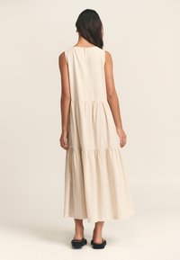 Ľahké béžové šaty bez rukávov s vrstvenou sukňou, jemnou textúrou a dĺžkou po kolená. Majú okrúhly výstrih a jemný detail s gombíkom na chrbte.