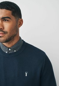 Granatowy sweter w prążki z teksturowanej tkaniny, z logo białego jelonka, założony na koszulę w kratę z kołnierzykiem w czerni i bieli.