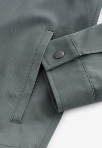 Gros plan d'une manche de veste en tissu gris-vert avec un poignet à bouton-pression et une poche passepoilée verticale à proximité.