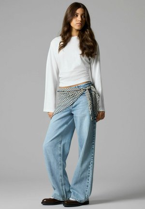 Mladá žena s dlhými vlnitými vlasmi, oblečená v bielom tričku s dlhým rukávom, svetlomodrých džínsoch s širokými nohavicami, hnedých topánkach a s viazaným šálom s bodkami okolo pása.