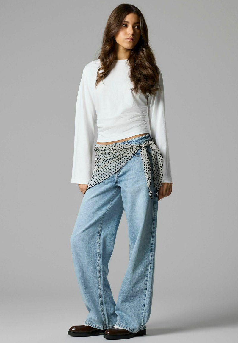 Jeune femme aux longs cheveux ondulés portant un haut blanc à manches longues, un jean large bleu clair, des chaussures marron et un foulard à pois noué autour de la taille.