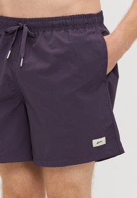 Męskie kąpielówki w jednolitym ciemnofioletowym kolorze, z elastycznym pasem ze sznurkiem, bocznymi kieszeniami i naszywką z logo na dolnym zakończeniu.