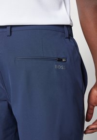Pantalons navy en tissu lisse, dotés d'une poche arrière zippée et de marquages discrets. Coupe ajustée avec des détails cousus.
