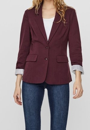 Mujer que lleva un blazer ajustado de color púrpura oscuro con puños a rayas remangados sobre una camiseta blanca y vaqueros azules, de pie frente a un fondo liso.