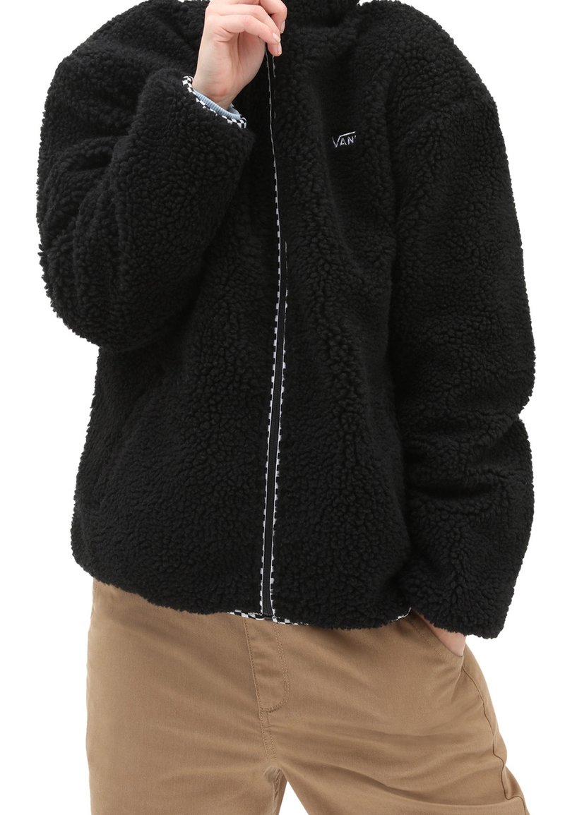 Personne portant une veste noire duveteuse zippée, une main dans la poche et l'autre touchant son visage, associée à un pantalon marron clair.