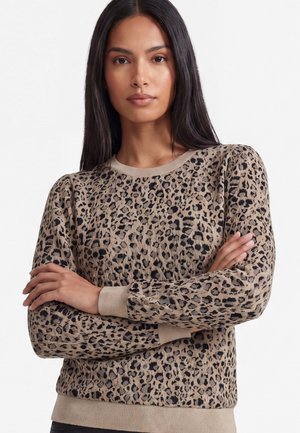 Donna con capelli lunghi e scuri che indossa un maglione beige e nero con stampa leopardata, in piedi con le braccia incrociate davanti a uno sfondo semplice.