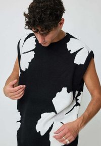 Pull en maille sans manches noir avec de grands motifs floraux blancs, col rond et bords côtelés. Le tissu semble doux et texturé.