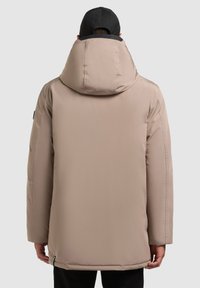 Chaqueta acolchada beige con una capucha grande, textura suave, bolsillos laterales y corte recto, llevada sobre pantalones negros. Gorra negra en la cabeza.