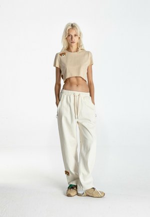 Mujer rubia de pie con las manos en los bolsillos, lleva un top corto beige, pantalones sueltos blancos con parches de ositos de peluche y zapatillas beige.