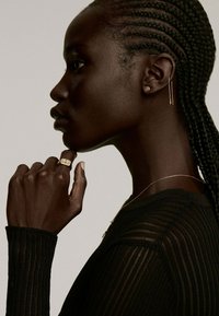 Joyería de oro en una mano y una oreja, llevada por una persona con el cabello trenzado, vestida con una camiseta de manga larga negra y texturizada, contra un fondo claro.