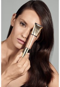 Estee Lauder Futurist Hydra Rescue fuktighetsgivende sminke i en rosa tube med gullakenter, holdt av en modell mot en grå bakgrunn.