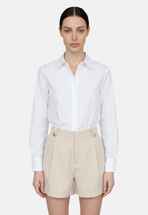 Donna con capelli scuri che indossa una camicia bianca con bottoni e pantaloncini beige a vita alta con bottoni dorati, in piedi di fronte su uno sfondo semplice.