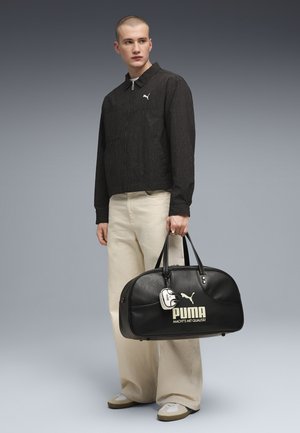 Jeune homme portant une veste noire zippée et un pantalon large crème, tenant un sac de voyage noir Puma avec une étiquette blanche sur un fond gris.