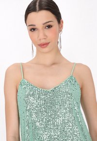 Camisole verde con paillettes, dotato di spalline sottili, caratterizzato da un collo a V e una consistenza liscia. Le paillettes argentate riflettono la luce, aggiungendo brillantezza.